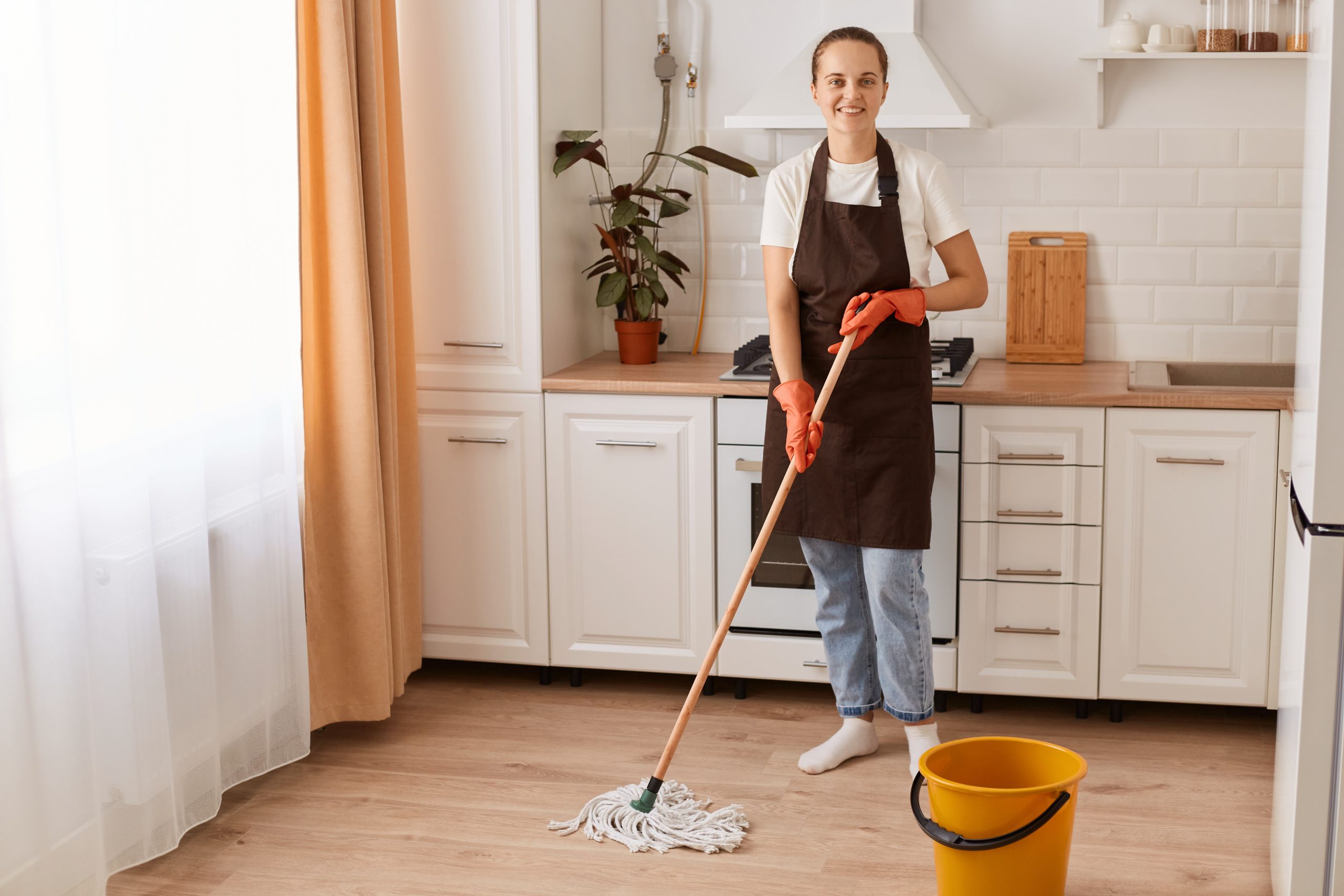 Full length portrait of attractive cheerful hardworking woman making fast domestic work, washing floor in modern light white interior kitchen, wearing brown apron, looking at camera.